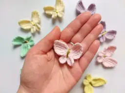 Różowy motylek na szydełku w dłoni, otoczony innymi kolorowymi motylkami.