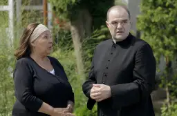 Kobieta w czarnej bluzce i mężczyzna w sutannie rozmawiają na tle zieleni.