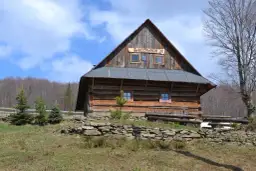 Drewniana bacówka w górach z napisem "BACÓWKA" na dachu, otoczona drzewami i kamiennym murkiem.