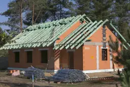 Budowa domu w metodzie gospodarczej: wzniesione ściany z pustaków ceramicznych, gotowy szkielet dachu z zielonych krokwi, materiały budowlane na placu.