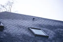 Dach pokryty ciemną dachówką betonową płaską, z widocznym oknem dachowym i kominkiem wentylacyjnym.