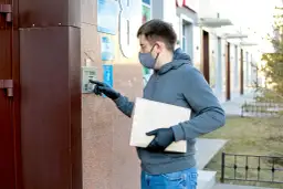 Mężczyzna w masce i rękawiczkach trzyma paczkę i wpisuje kod do domofonu, aby dostarczyć przesyłkę.