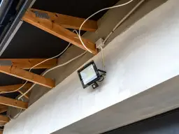 Nowoczesna elektryka w garażu: jasny reflektor LED z czujnikiem ruchu zamontowany na betonowej belce, otoczony drewnianą konstrukcją dachu i białymi przewodami.