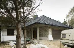 Budowa domu w toku. Widać białe płyty styroduru przy ścianach, które będą izolacją. Obok stoi przyczepa kempingowa.