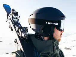 Narciarz z nartami na ramieniu, w czarnym kasku HEAD i goglach, gotowy na górskie szaleństwo.