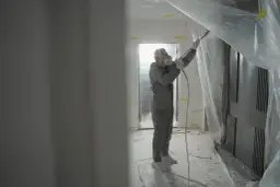 Pracownik w kombinezonie i masce maluje ściany agregatem. Świetny pomysł na ściany bez gładzi, tworzy nowoczesny efekt.