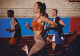 Trzech biegaczy na bieżni. Kobieta w pomarańczowym topie i beżowych legginsach, mężczyzna w beżowej koszulce i pomarańczowych spodenkach, oraz mężczyzna w białej koszulce i białych spodenkach. To jest ich "drop" formy.