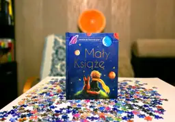 Odkryj, dlaczego warto przeczytać Małego Księcia. Książka z liskiem i chłopcem na tle kosmosu, otoczona rozsypanymi puzzlami.