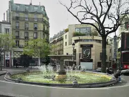 Fontanna na placu Pigalle w Paryżu, otoczona kamienicami i drzewem.