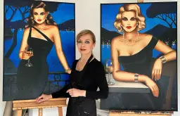 Künstlerin steht zwischen zwei Gemälden von eleganten Frauen in schwarzen Kleidern mit Gläsern Wein.