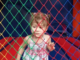 Mała dziewczynka w kratkowanej sukience patrzy przez kolorową siatkę. Może słucha piosenki dla małych dzieci.