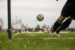 Piłkarz kopie piłkę na boisku, w tle inni zawodnicy i bramka.