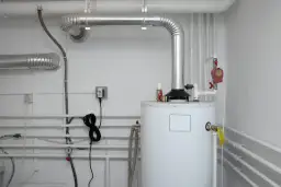 Biały bojler CO z metalowymi rurami i instalacją hydrauliczną na białej ścianie.