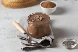 Słoik z czekoladowym puddingiem proteinowym, posypanym wiórkami czekolady, obok łyżeczki i miseczki z nasionami chia.