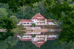 Biały hotel z czerwonym dachem nad spokojnym jeziorem, odbijający się w wodzie.
