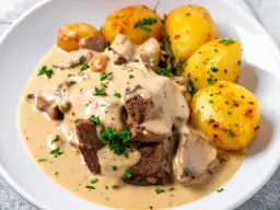 Schab w kremowym sosie z grzybami i pieczonymi ziemniakami, udekorowany natką pietruszki.