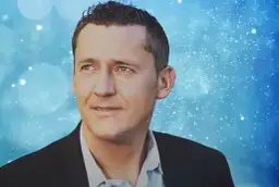 Portret Adama Chroli w garniturze na tle niebieskich, migoczących świateł.