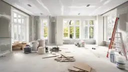 Salon w trakcie remontu, widoczne materiały budowlane, drabina i okna z widokiem na drzewa.