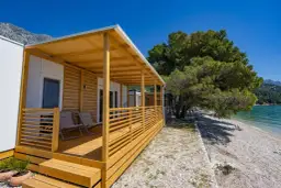 Domek mobilny z drewnianym tarasem i widokiem na morze w camp Dole Živogošće. Dwa leżaki czekają na plaży.