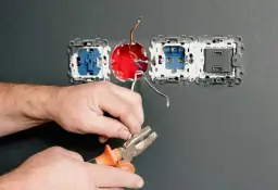 Ręce majsterkowicza przygotowują przewody do podłączenia kilku gniazdek elektrycznych. Narzędzia i instalacja na szarej ścianie.