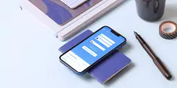 Smartfon z aplikacją do wymiany walut HKD na PLN, obok pióro i notatniki.