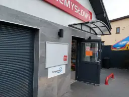 Wejście do sklepu z otwartymi drzwiami, tablicą ogłoszeń i logo "Orlen paczka".