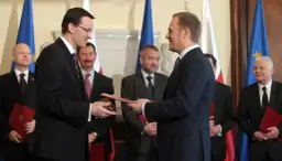 Mateusz Morawiecki przekazuje dokument Donaldowi Tuskowi, stojąc przed polskimi i unijnymi flagami.