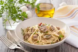 Talerz spaghetti z pieczarkami, posypany parmezanem i natką pietruszki. Obok sztućce, oliwa i zioła.