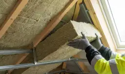 Ręce w rękawiczkach montują wełnę mineralną, docieplenie dachu. Sprawdź, jakie czeka Cię ocieplenie dachu dofinansowanie.