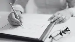 Dłoń pisze na dokumencie, obok okulary. Dowiedz się, jak sprawdzić pozwolenie na budowę sąsiada, analizując formalności.