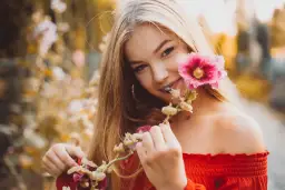 Uśmiechnięta blondynka w czerwonej bluzce podczas sesji kobiecej w plenerze, trzymająca gałązkę z różowymi kwiatami malwy.