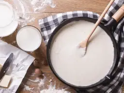 Gładki, biały sos w patelni, obok mleko, masło i gałka muszkatołowa.