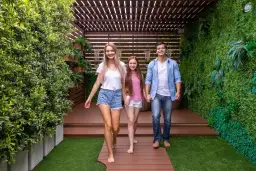 Rodzina idzie po drewnianej ścieżce na tarasie z zieloną ścianą i pergolą.