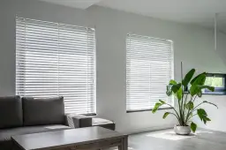 Nowoczesne wnętrze z szarą kanapą i stolikiem. Jasne, białe żaluzje drewniane na oknach tworzą ciekawe wzory światła na podłodze.