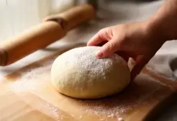 Dłoń dotyka kulki ciasta na pizzę posypanej mąką na desce, obok wałek.