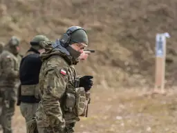 Żołnierz w mundurze maskującym, słuchawkach i okularach ochronnych celuje z pistoletu na strzelnicy.