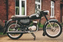 Czarny motocykl WSK 125, z charakterystycznym emblematem na baku, który sugeruje jego pojemność. Stoi na bruku przed ceglanym budynkiem.