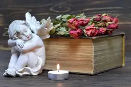 Figurka aniołka, stara księga, świeca i uschnięte róże na drewnianym tle.