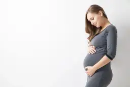 Uśmiechnięta ciężarna kobieta w szarym ubraniu delikatnie dotyka brzucha. To naturalne zmiany, jak rozstępy w ciąży, które są częścią tej wyjątkowej podróży.