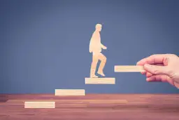 Drewniana postać człowieka wchodzi po schodkach, a dłoń dokłada kolejny stopień.