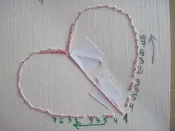 Instrukcja jak wyhaftować serce: różowa nić przechodzi przez dziurki, tworząc kontur serca. Liczby wskazują kolejność.