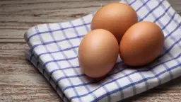 Trzy brązowe jajka na niebiesko-białej kratkowanej ściereczce na drewnianym tle.