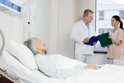 Starsza pacjentka w łóżku szpitalnym, lekarz i pielęgniarka omawiają jej stan, potencjalne czynniki jatrogenne.