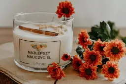 Świeca w szklanym naczyniu z napisem "Najlepszy Nauczyciel" i pomarańczowe kwiaty.