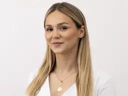 Kobieta z blond włosami i delikatnym makijażem, uśmiecha się lekko, choć jej nienaturalny wyraz twarzy sugeruje, że pozuje do zdjęcia.