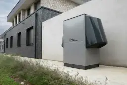 Nowoczesna pompa ciepła, szara, z logo "alpha innotec", stoi przy betonowej ścianie obok budynku z cegły.