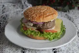 Burger z sezamową bułką, kotletem, serem, pomidorem, sałatą i cebulą na białym talerzu.