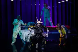 Na scenie, w surrealistycznej atmosferze, panna obsada w stroju z głową kozła siedzi na wózku. Wokół niej, w jasnych fartuchach, poruszają się postacie przypominające personel medyczny.