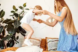 Radosne zabawy z 3 latkiem w domu. Mama trzyma za ręce skaczące dziecko na łóżku, otoczeni roślinami i poduszkami.