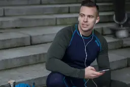 Mężczyzna w zielonej koszulce termoaktywnej z granatowymi rękawami i niebieskimi szwami, słucha muzyki na telefonie, siedząc na schodach.